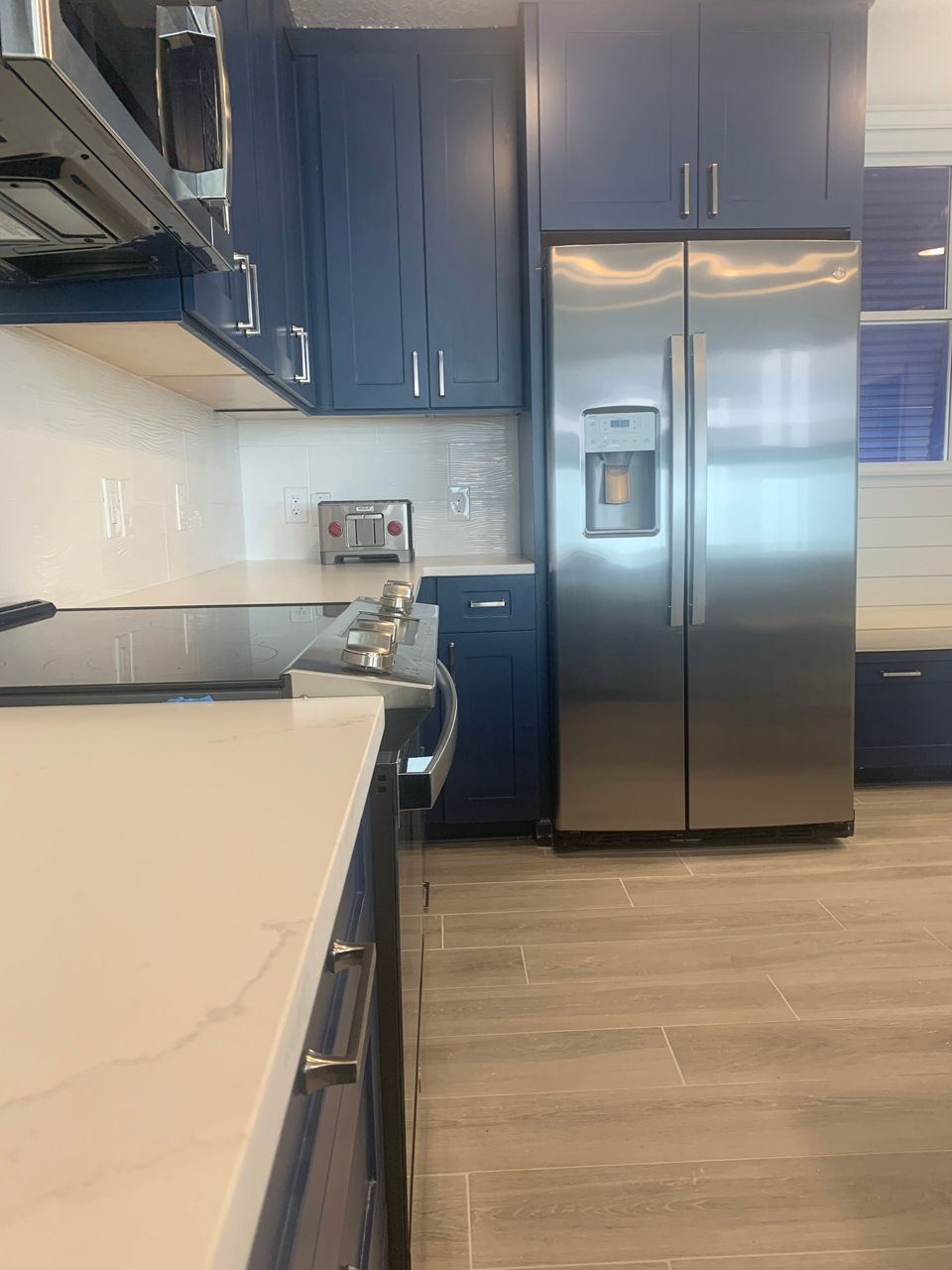Blue kitchen cabinets with white countertops, stainless steel refrigerator, and beige tiled floor.