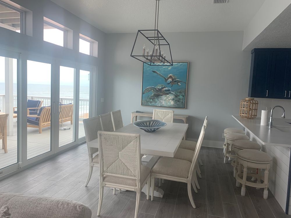 Dining room with water views; white table, chairs, art; blue-grey walls and hardwood floors.