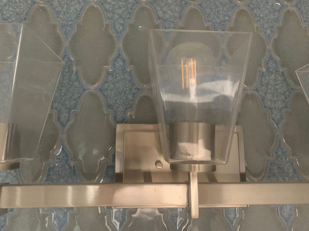 Brushed nickel vanity light fixture with a clear glass shade, mounted on blue patterned tile.