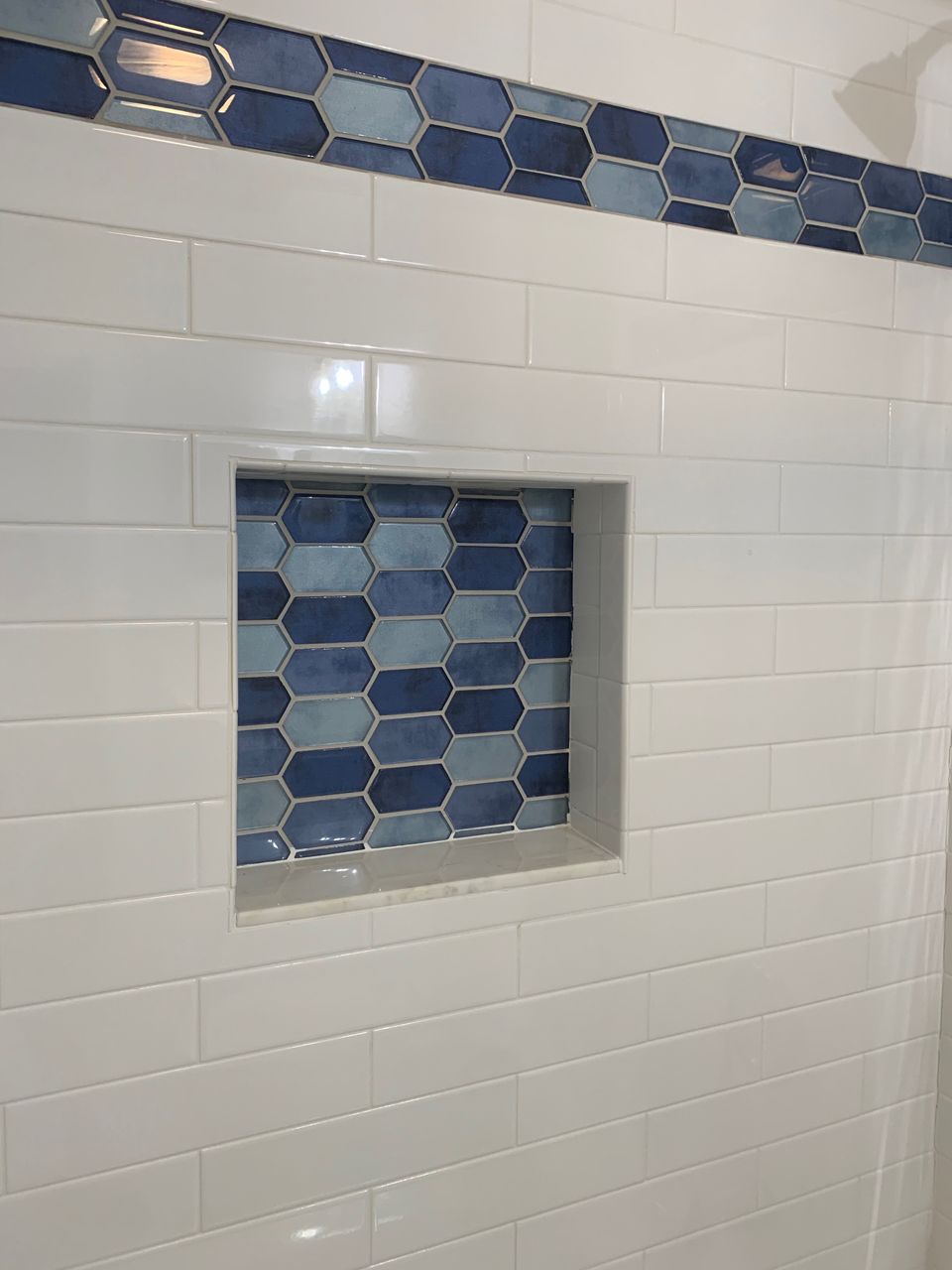 White tiled shower wall with blue hexagon accent tiles, recessed shelf with blue-and-white tiled back.