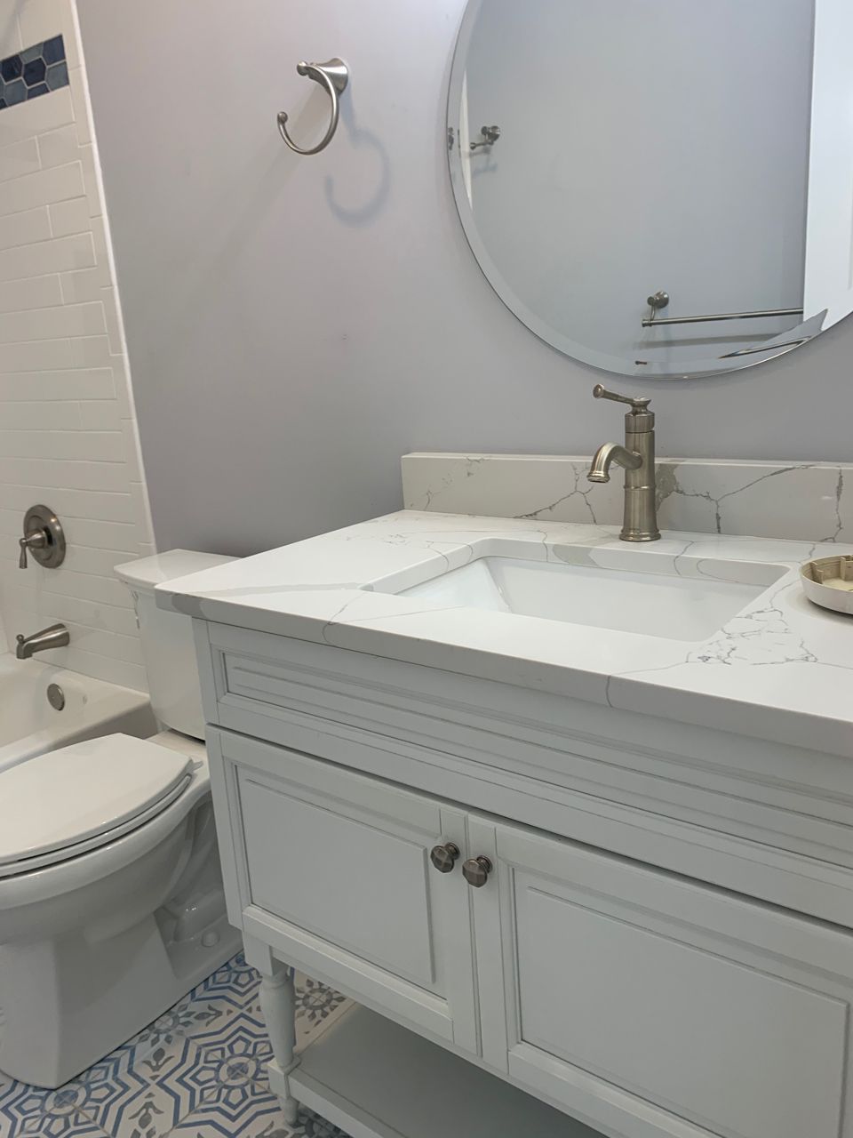 White bathroom vanity with marble countertop and sink, round mirror, toilet, and patterned tile floor.