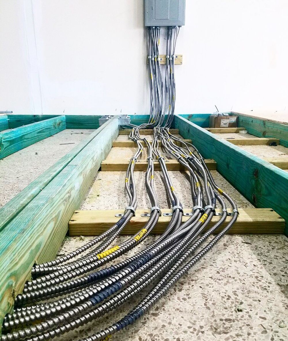 Electrical conduit cables running along wooden floor beams towards a gray electrical panel on a white wall.