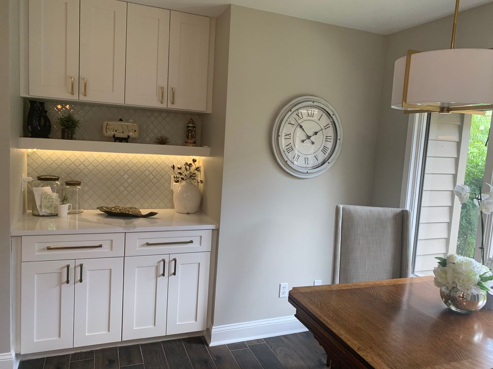 White built-in cabinet with decor, a clock on a beige wall, and a dining table near a window.