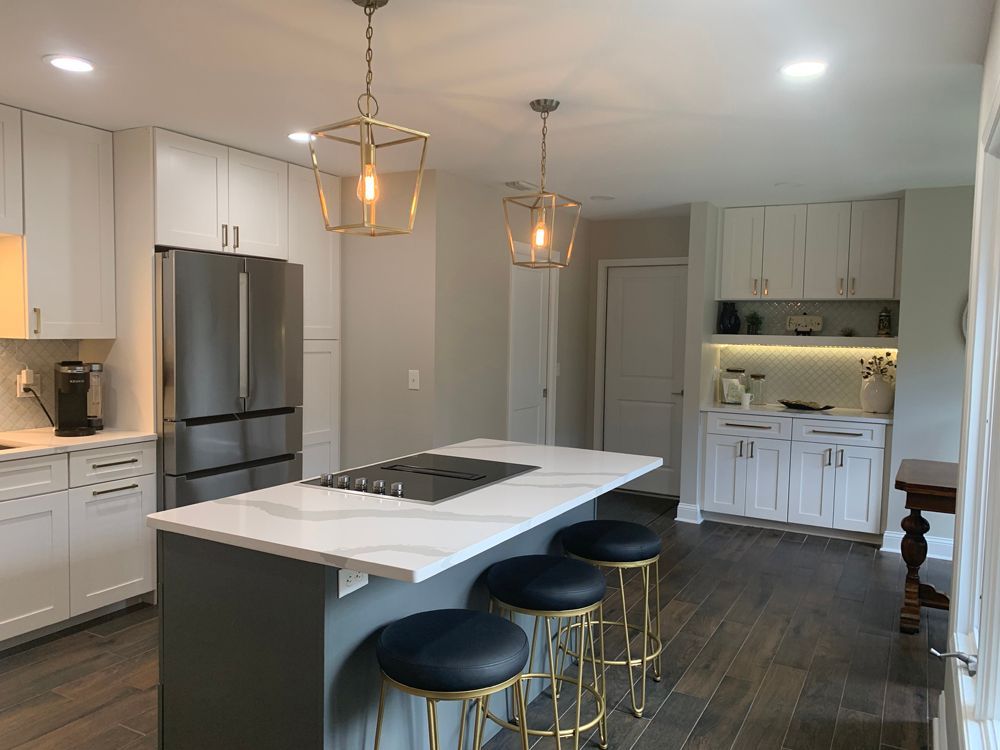 Modern white kitchen with island, stainless steel appliances, gray cabinets, gold light fixtures, and blue bar stools.
