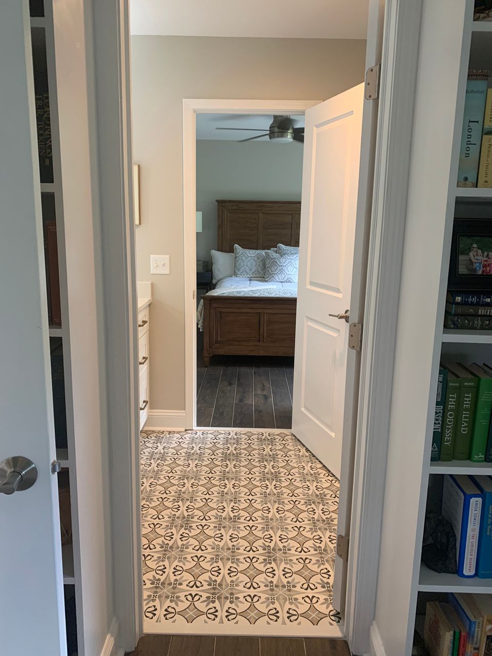 Hallway view into a bedroom, with patterned tile flooring and a bed visible. White walls, door frames, and bookshelves.