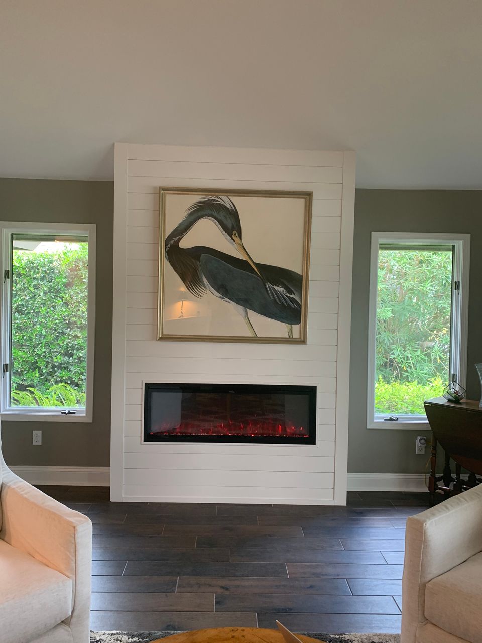 White fireplace with framed heron art, flanked by windows. Dark wood floor, light gray walls.