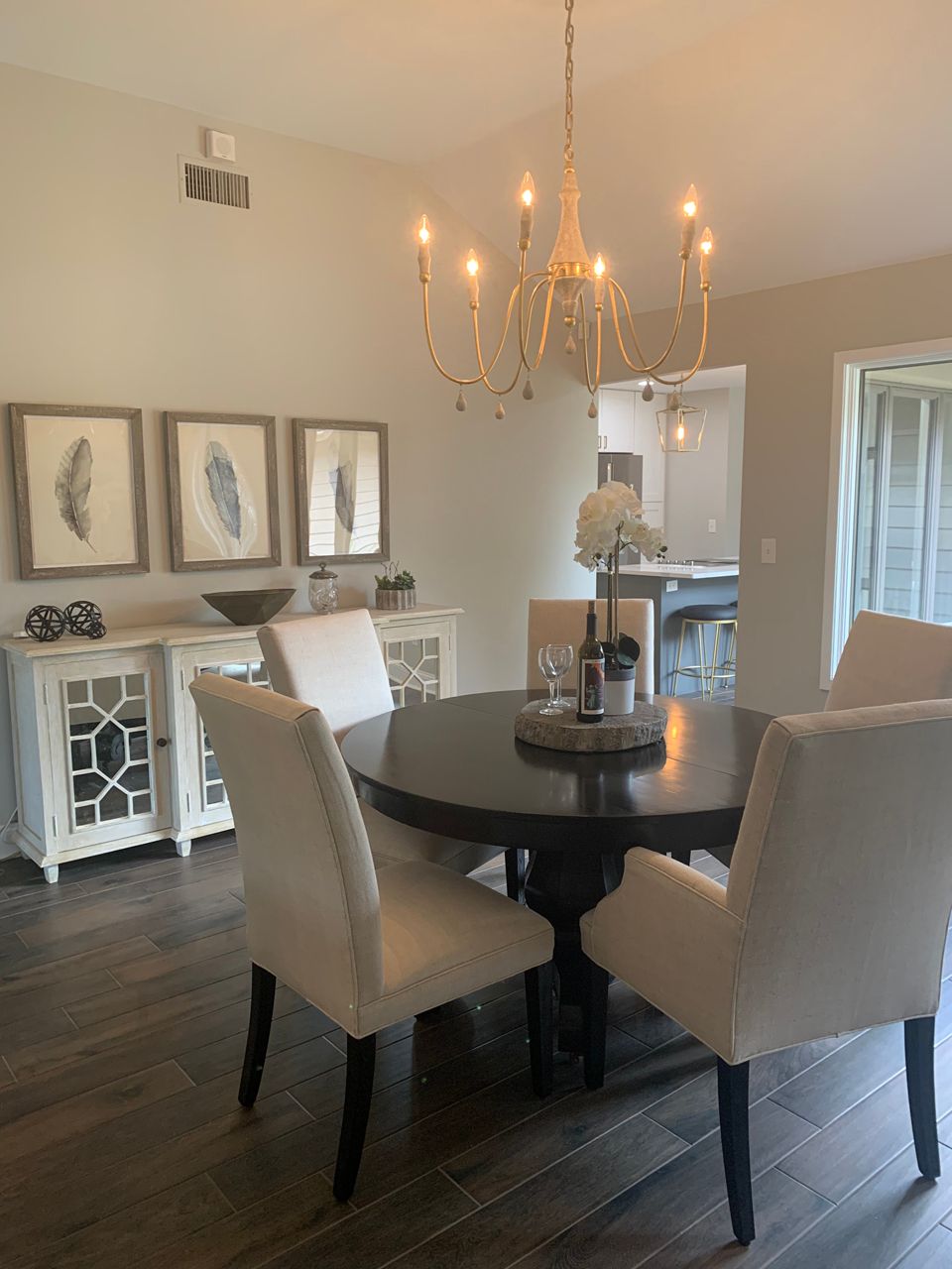 Dining room with round table, white chairs, sideboard, artwork, chandelier, and dark wood floors.