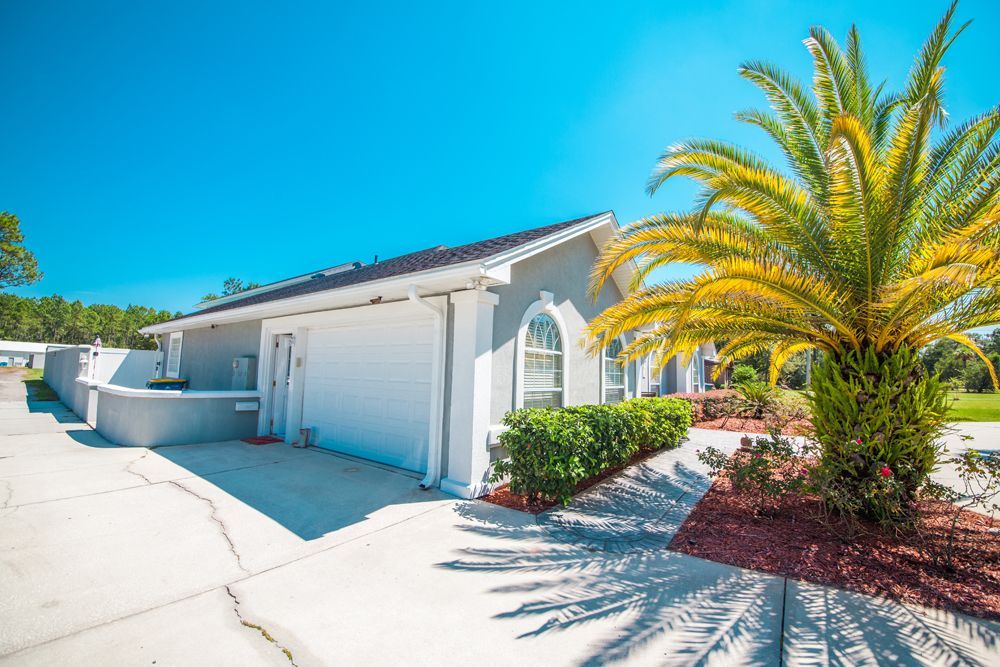 Exterior view of a light gray building with white garage doors, palm tree, and clear blue sky.