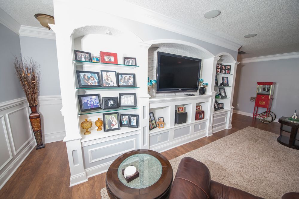 Living room with built-in white entertainment center, TV, shelves with photos, brown rug, and a round coffee table.