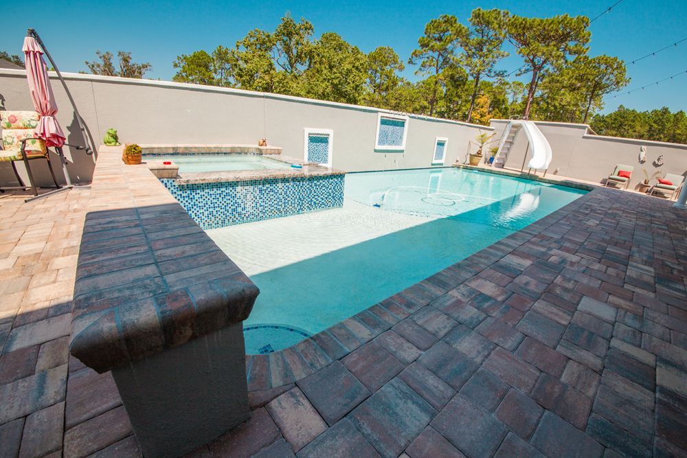 Backyard pool with blue water, tiled hot tub, stone deck, and gray wall.