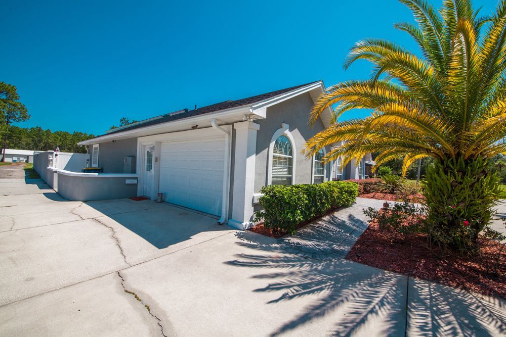 Gray stucco building with white garage door, arched window, and palm tree under a clear blue sky.