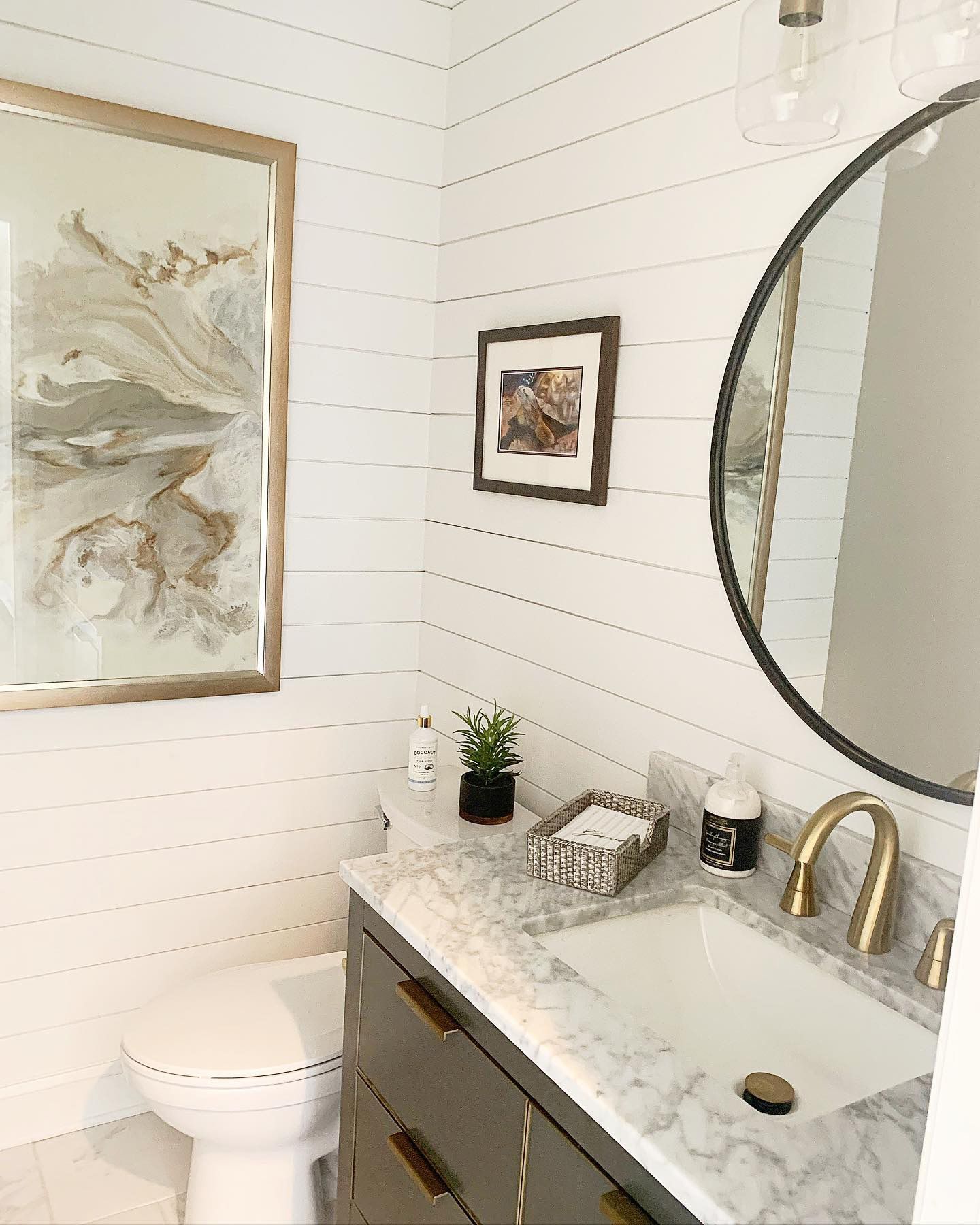 White-paneled bathroom with dark gray vanity, gold accents, round mirror, and two framed artworks.