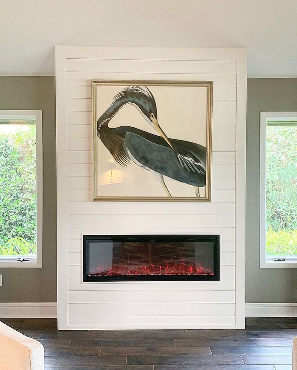 White shiplap fireplace with framed heron art above a glowing electric fire, flanked by windows.