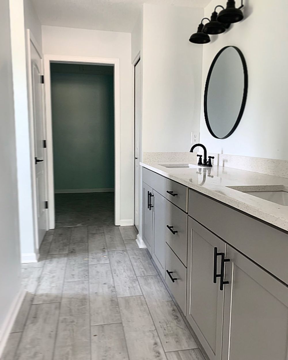 Bathroom with gray cabinets, countertops, black fixtures, oval mirror, and hallway with blue-green wall.