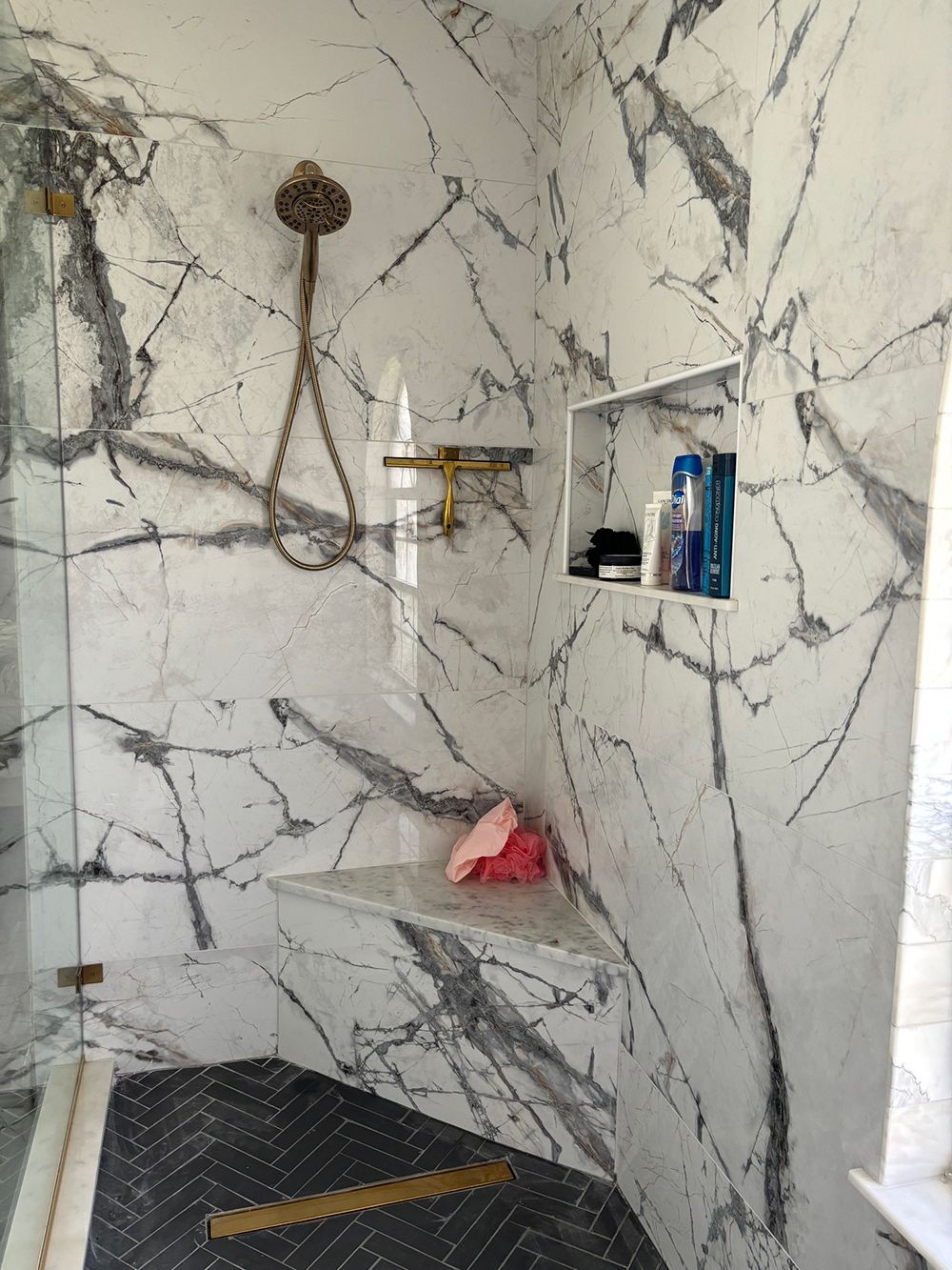 Marble-tiled shower with gray veining, gold fixtures, glass door, and a built-in shelf with toiletries.