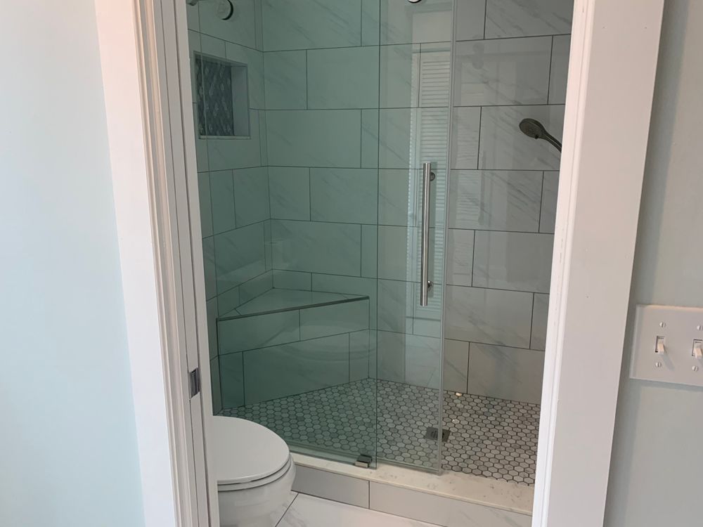 Modern shower with glass door, white tile walls, gray floor, and a toilet in a bathroom.
