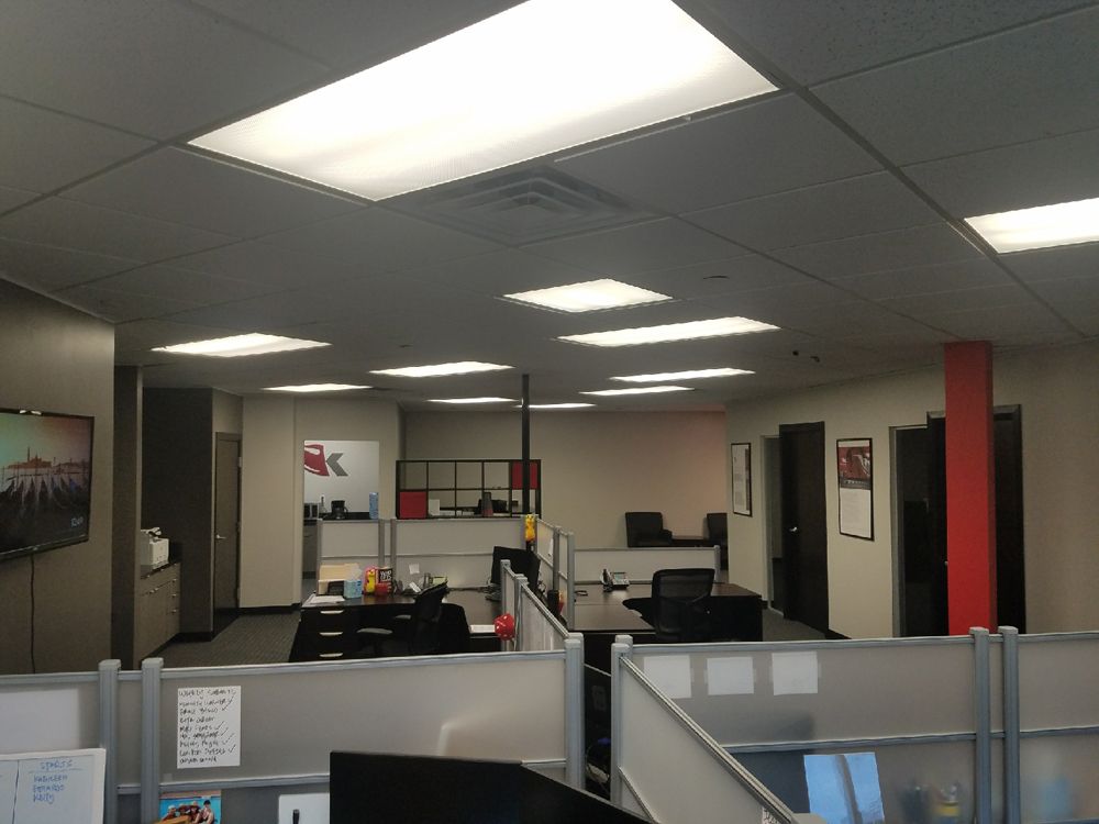 Interior view of an office with cubicles, desks, and overhead lighting; doors and windows are visible.