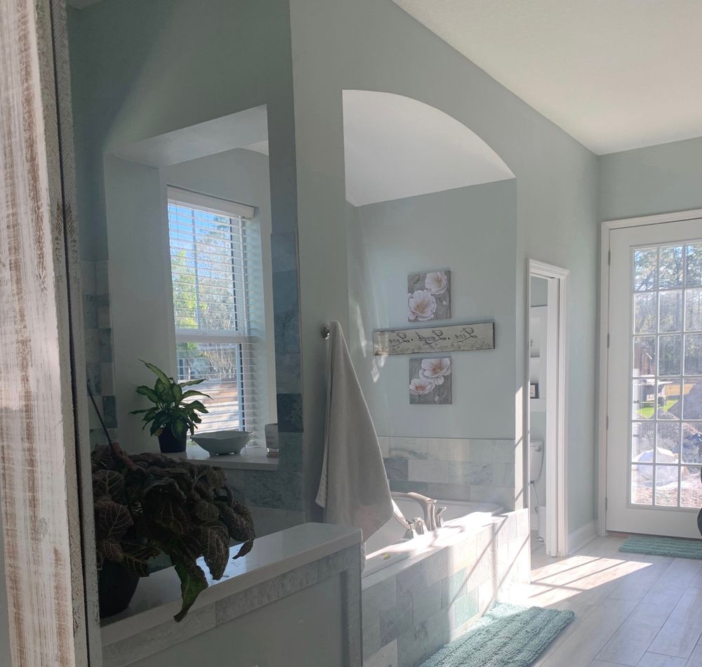 Bathroom with a window and tub. Light blue walls, white trim, green rug. Sunlight streams through.