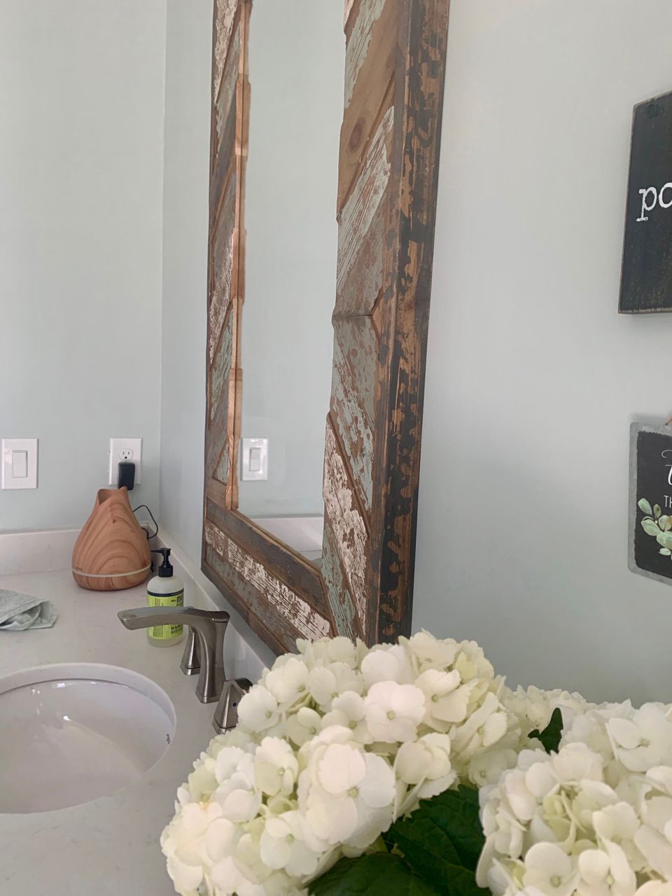 Bathroom with large wooden-framed mirror over a white sink, white hydrangeas, light blue walls.