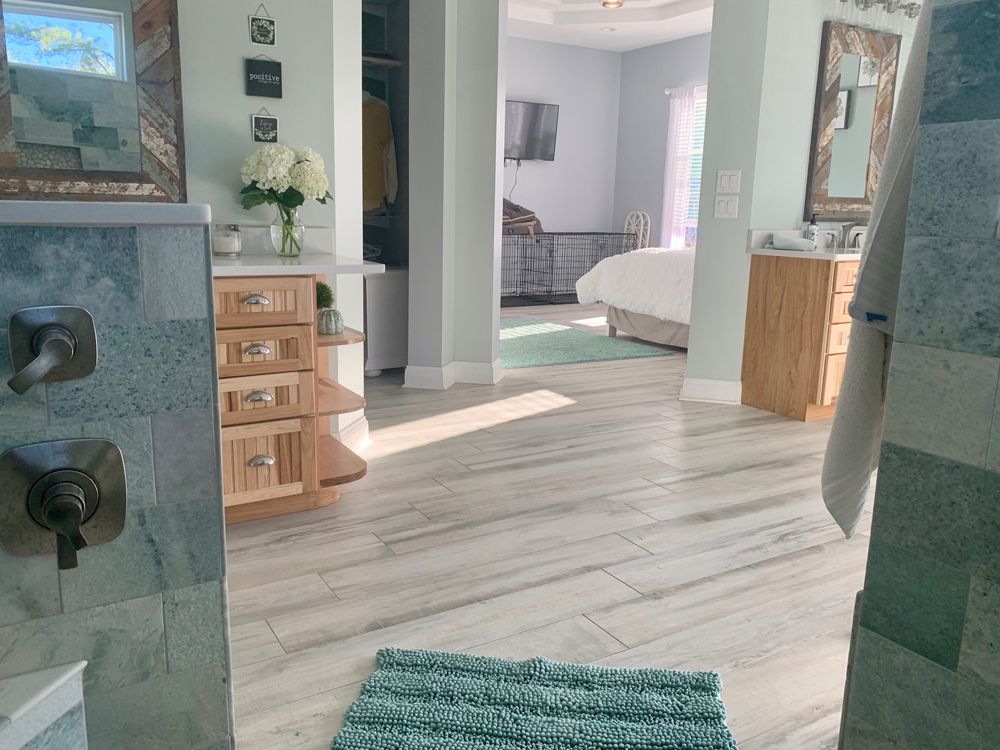 A light-filled bathroom with a view into a bedroom, featuring wood cabinets, marble floors, and a green rug.