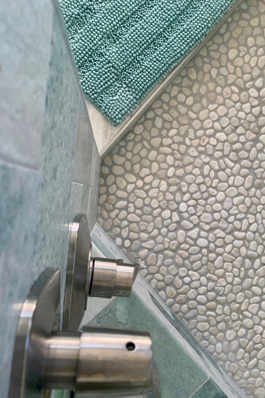 Close-up of stainless steel door handles on a teal tiled surface next to a pebbled floor with a teal bath mat.
