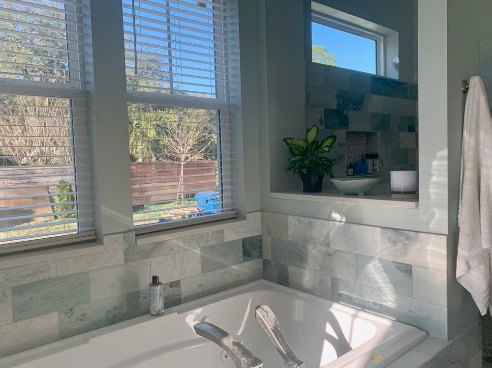 Bathroom with a tub, window with blinds, green and white tiled walls, and a small plant.