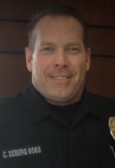 A police officer named C. Deberg, badge 6065, wearing a dark uniform, smiling at the camera against a brown background.