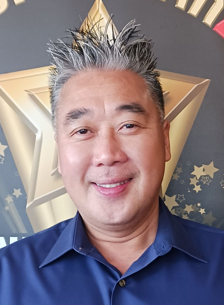 Smiling man with spiky gray hair, wearing a blue shirt, in front of a golden star.