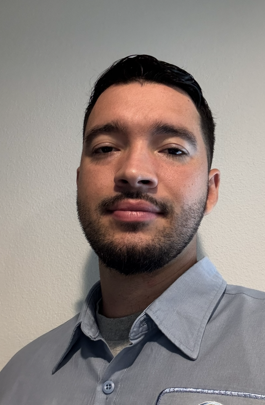 Gregory McGough Man with dark hair and a beard, wearing a light blue collared shirt, smiles gently.