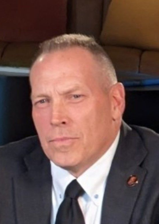 A man with short hair wearing a dark suit, white shirt, and black tie, positioned against a blurred background.