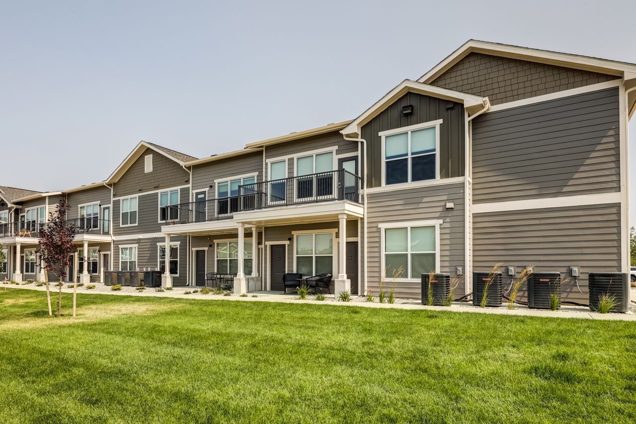 Row of modern gray townhouses with balconies, surrounded by green grass and a sunny sky.