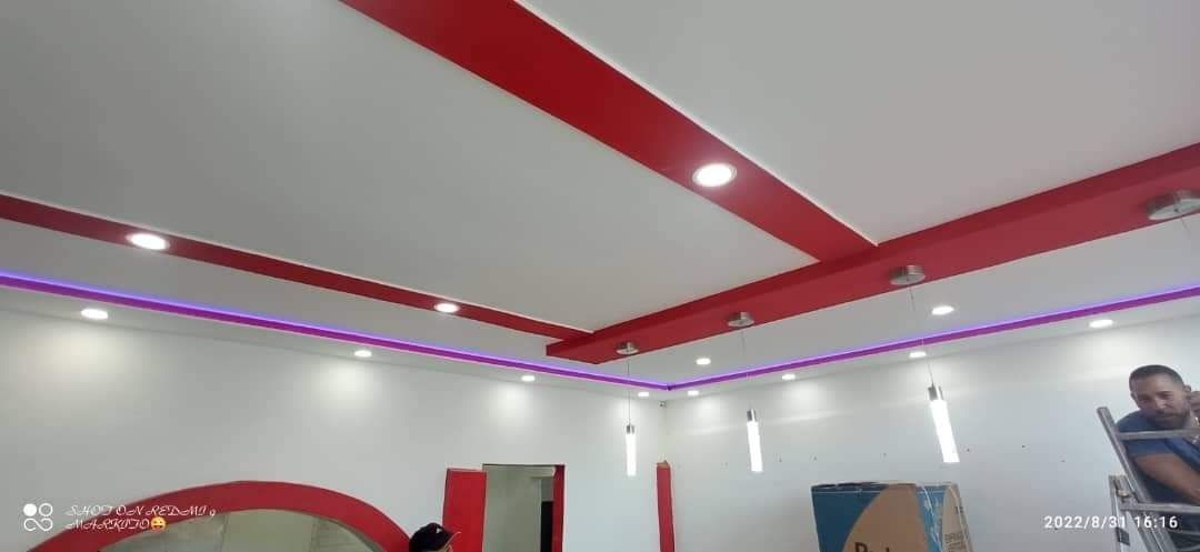 Habitación moderna con techo blanco, molduras rojas, tiras de luces LED moradas y una persona de pie a la derecha.