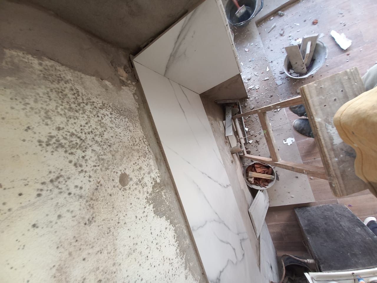 Primer plano de una esquina con azulejos blancos sucios, escombros y el borde de una silla de madera cerca.