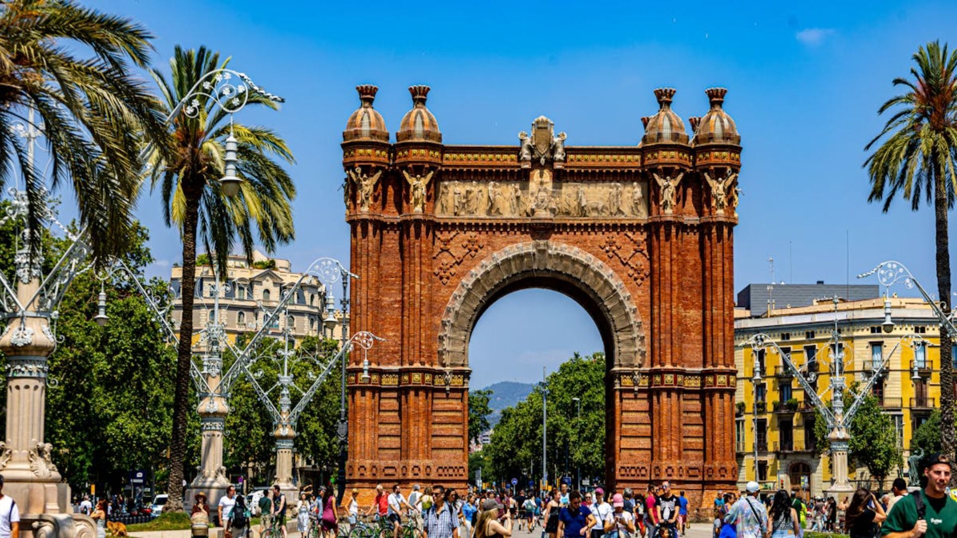 Arco ornamentado de ladrillo rojo en una plaza soleada con palmeras y una multitud al frente