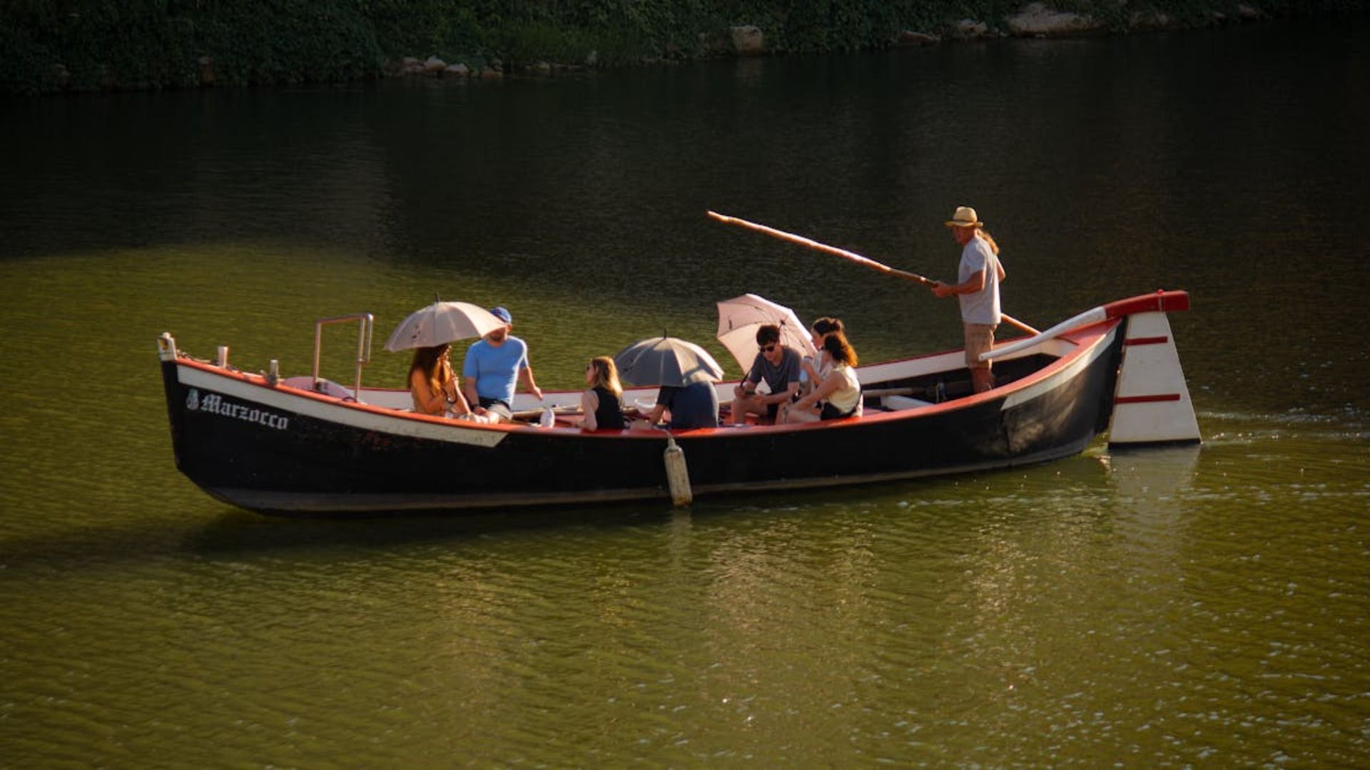 Personas en un bote negro en aguas tranquilas, una de ellas de pie con una caña de pescar cerca de una orilla boscosa.