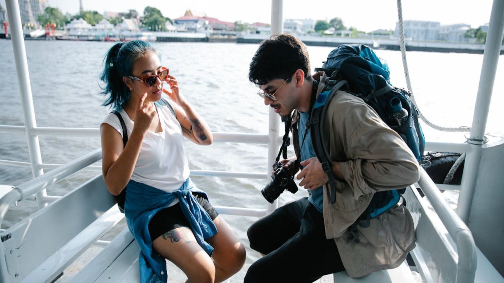 Dos personas sentadas en un bote, con mochilas y gafas de sol, charlando sobre el agua.
