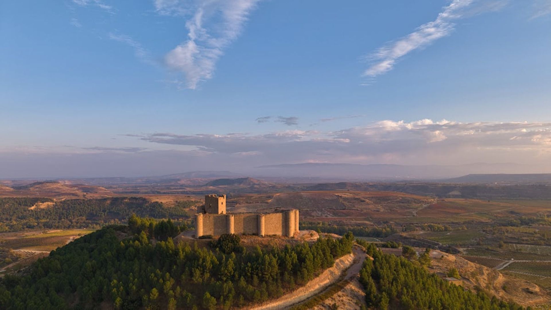 Fortaleza de piedra bañada por el sol en la cima de una colina verde con vistas a un amplio valle bajo un cielo azul.