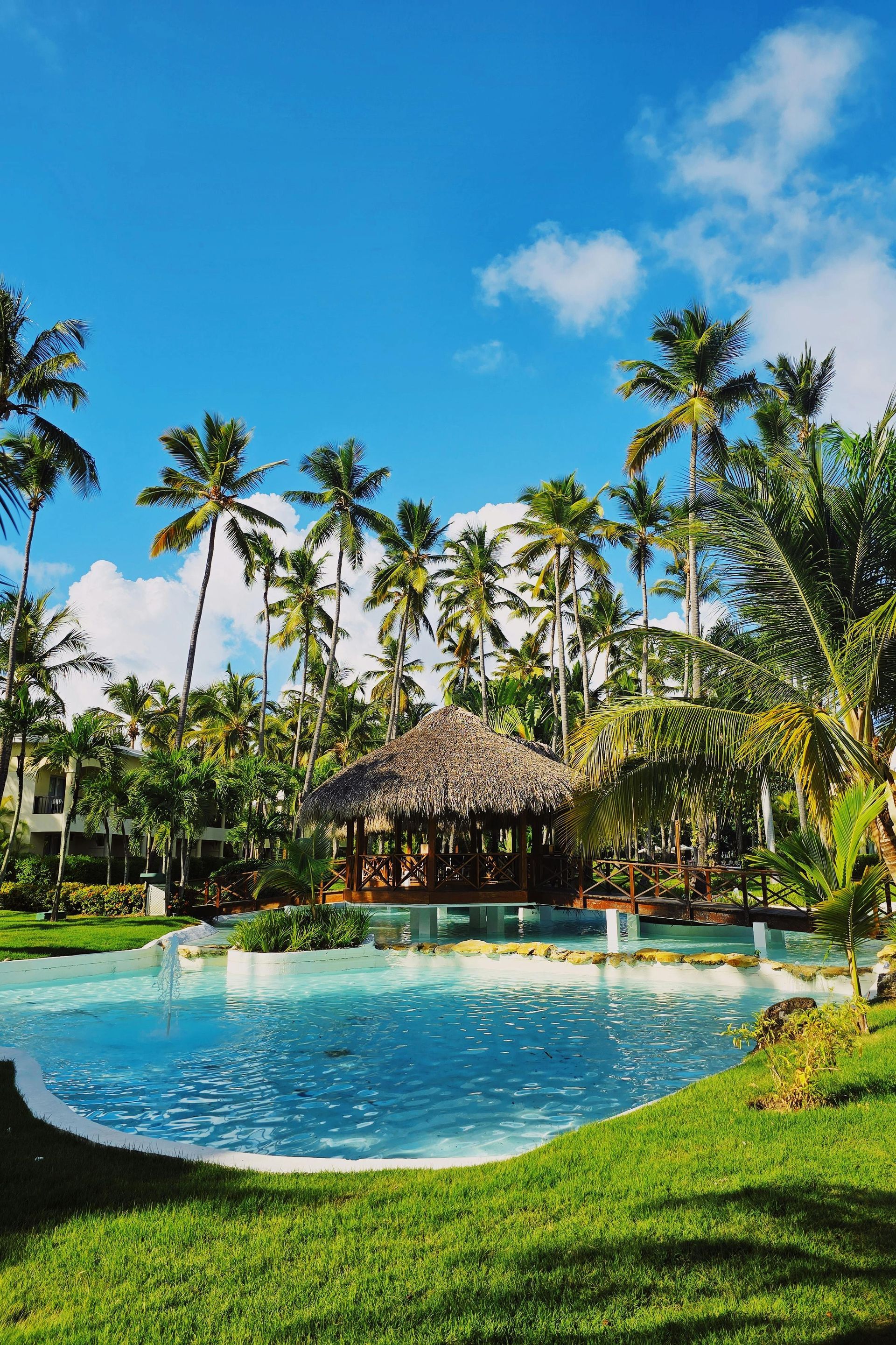 Piscina de resort tropical con agua turquesa, cabaña de paja, palmeras y cielo azul brillante.