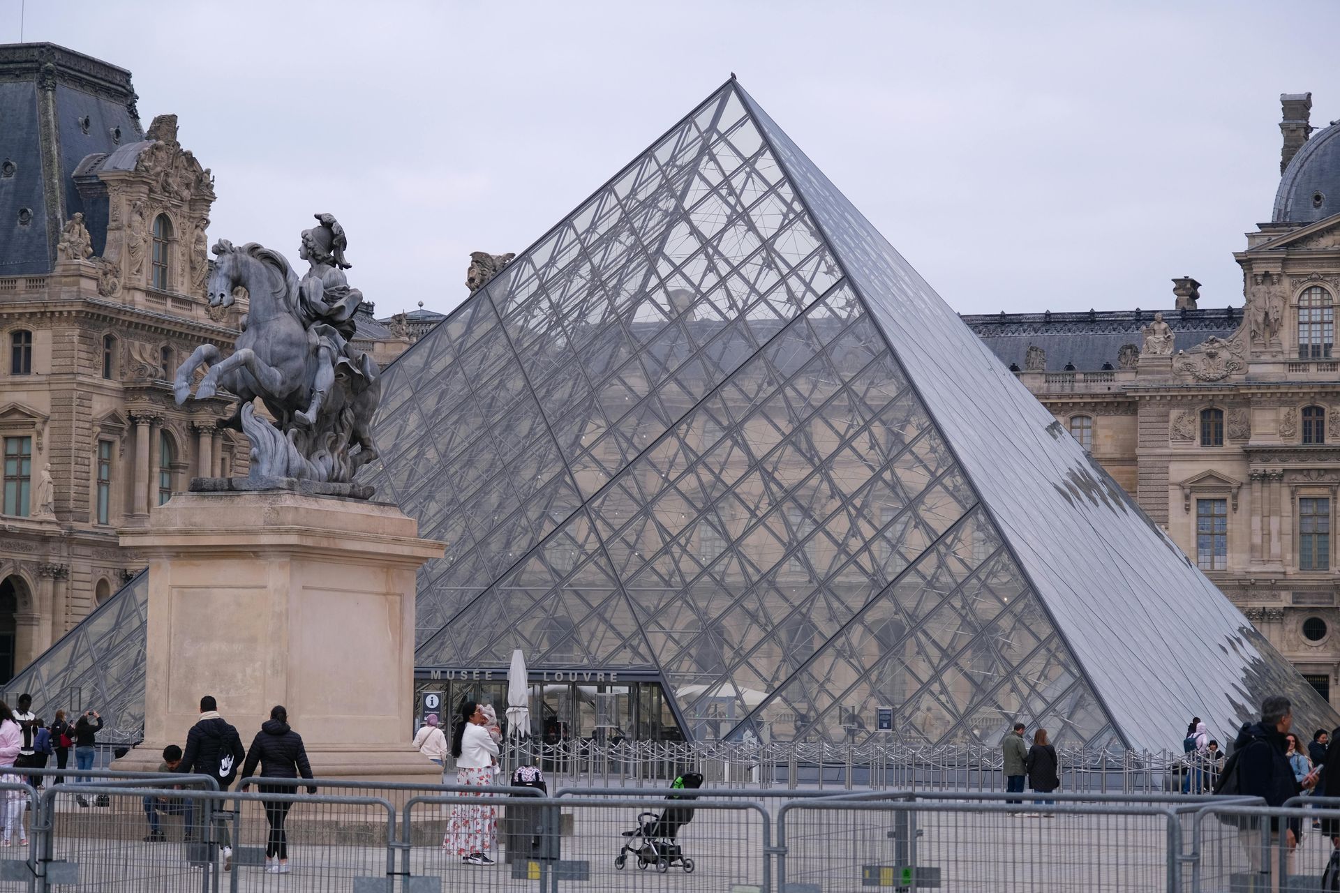Pirámide del Louvre en el patio de París con edificios históricos del museo y una estatua ecuestre de bronce.