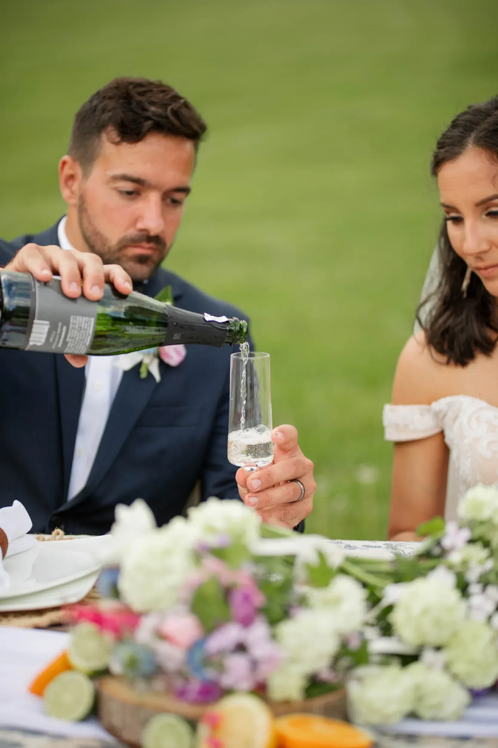 A bride and groom are sitting at a table while a man pours champagne into a glass.