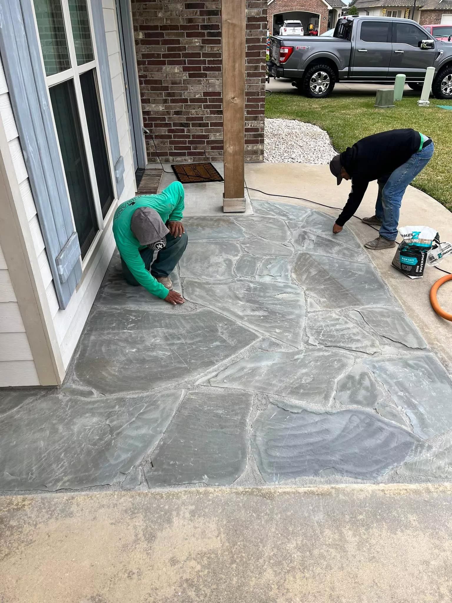 Two men are working on a stone patio in front of a house.