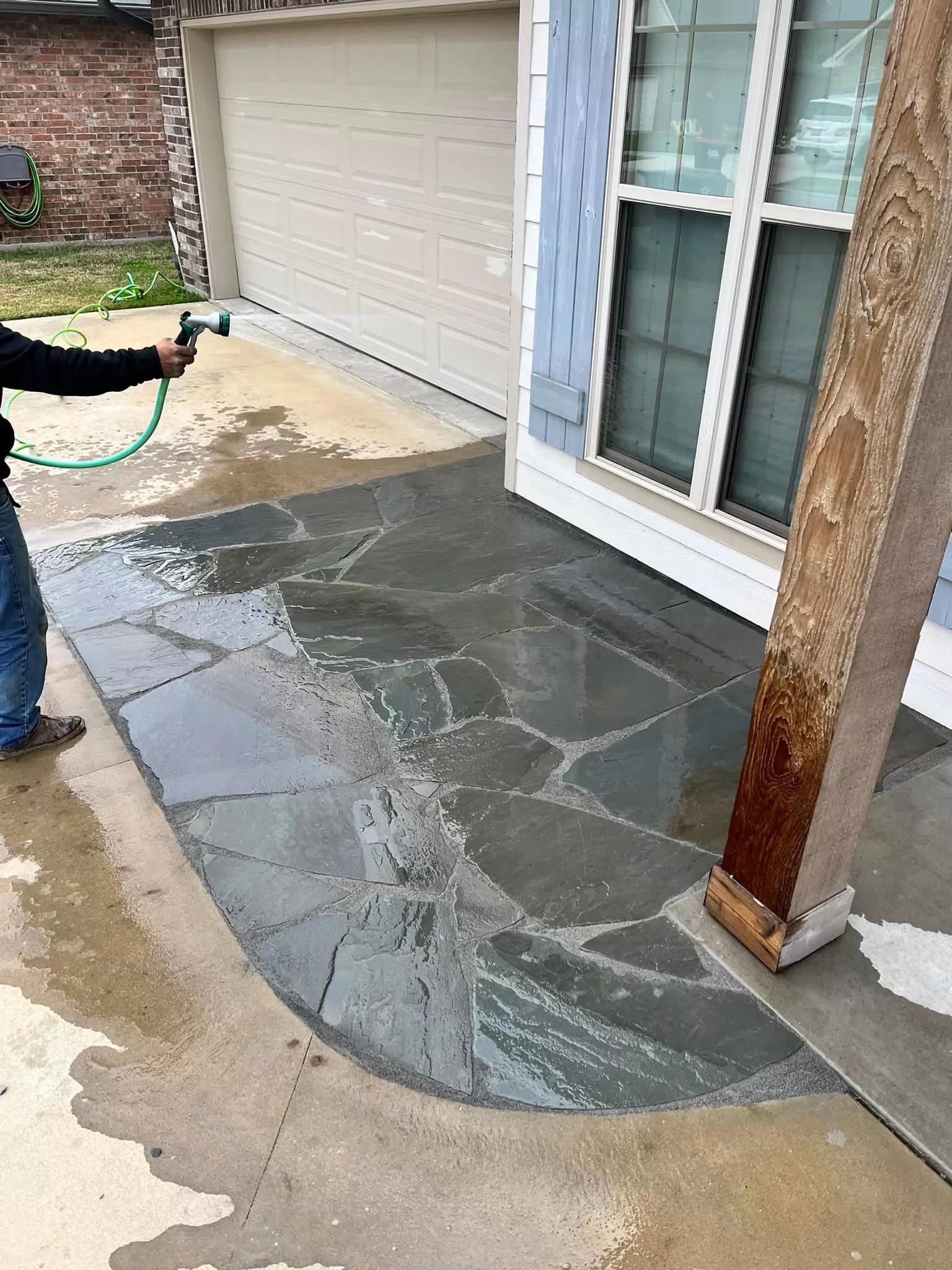 A man is holding a hose and spraying water on a stone walkway.
