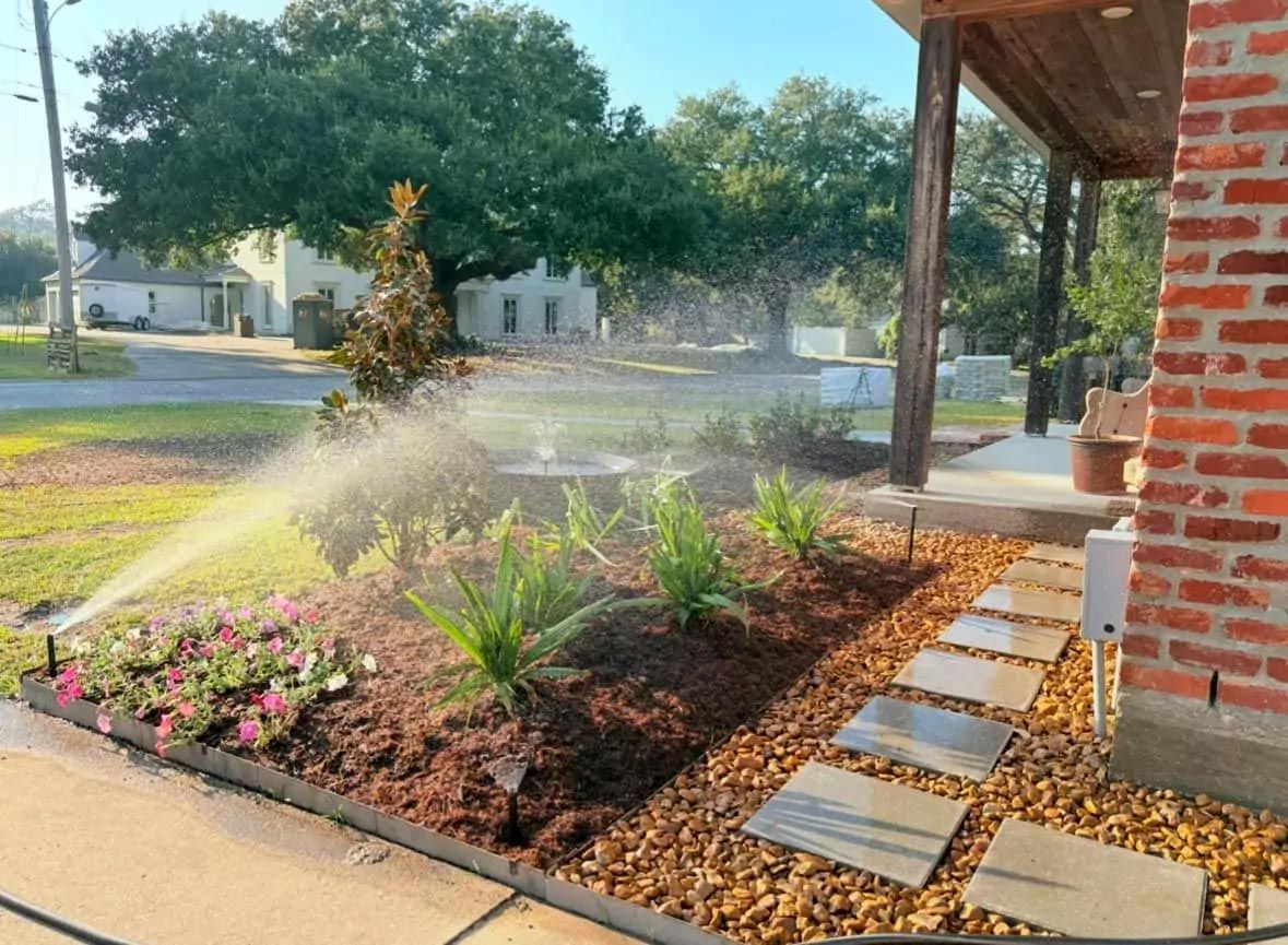 A sprinkler is spraying water in a garden in front of a brick building.