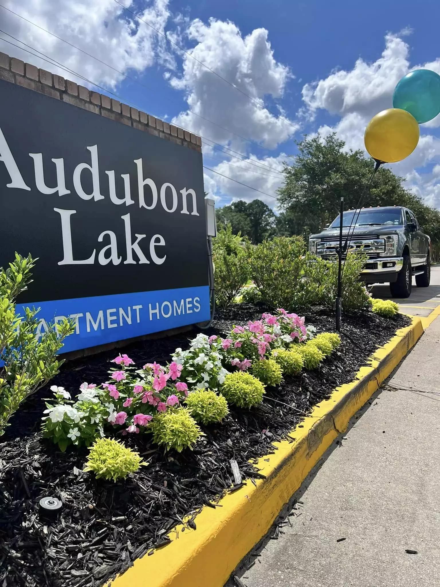 A sign for audubon lake apartment homes is surrounded by flowers and balloons.