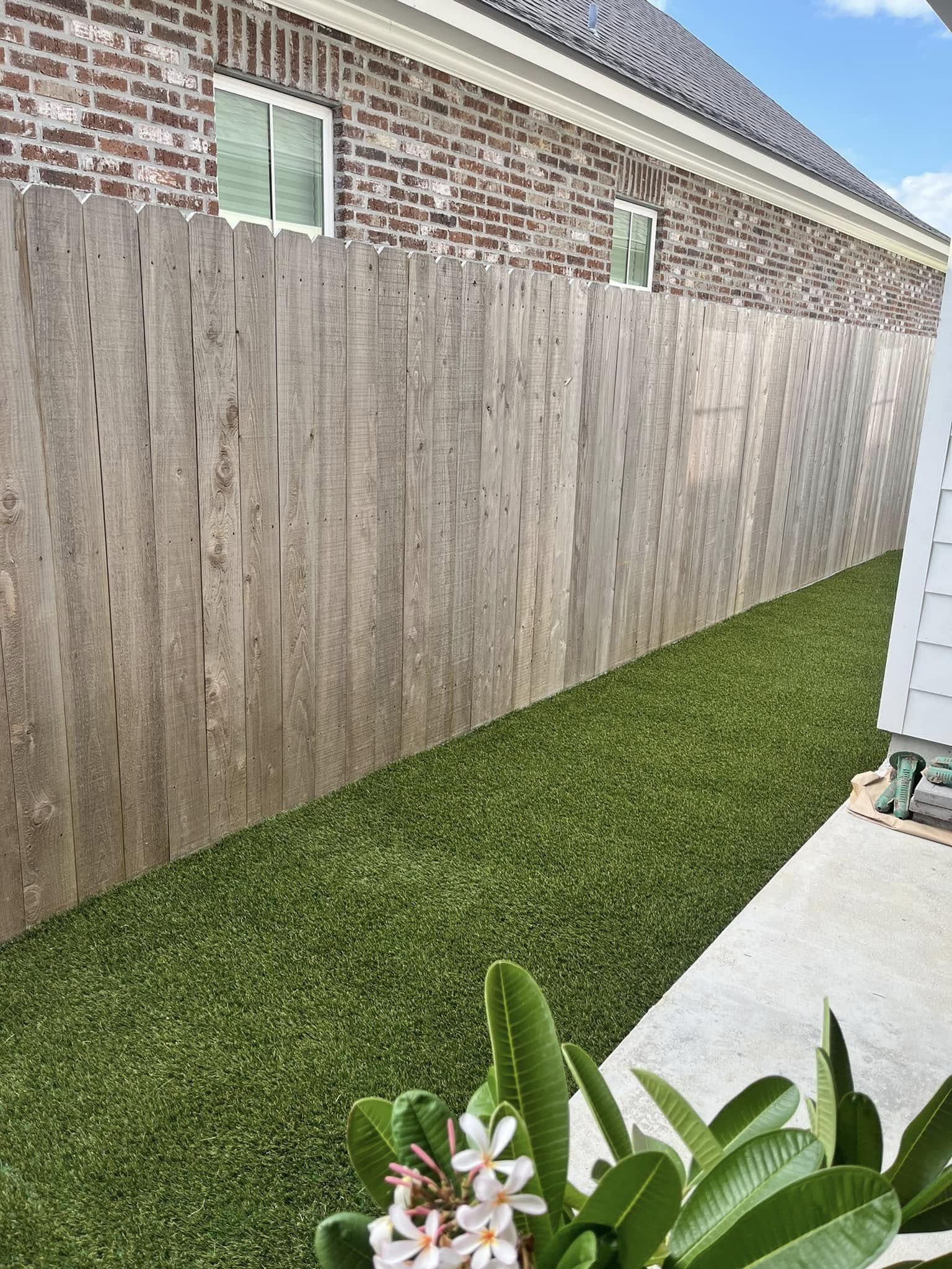 A wooden fence surrounds a lush green lawn in front of a brick house.