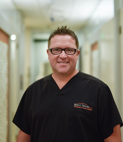 A man wearing glasses and a black scrub top stands in a hallway