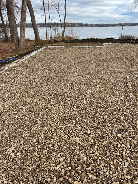 A gravel driveway next to a lake with trees in the background.