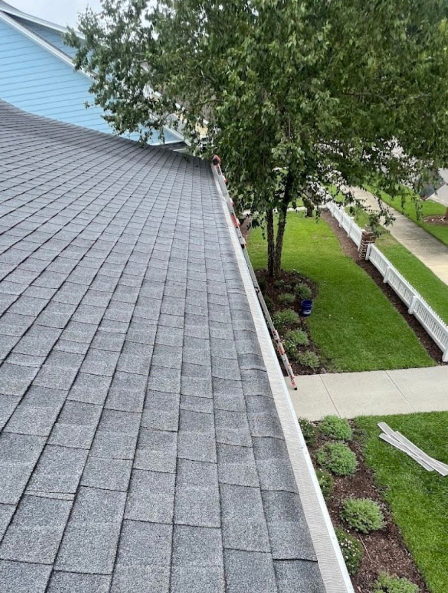 Gray shingle roof edge with a gutter and a tree next to a grassy lawn and sidewalk.