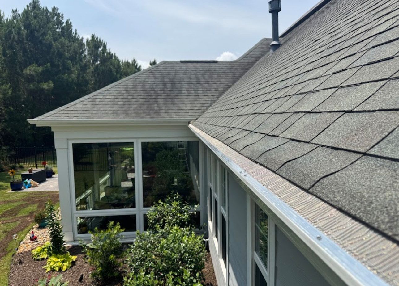 Gray shingled roof and gutter of a light-colored house with large windows; green plants in front.