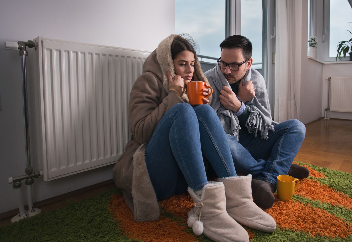 Young couple in jacket and covered with blanket sitting on floor beside radiator and trying to warm